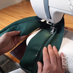 May include: A sewing machine stitching a dark green zipper onto fabric. The fabric is held in place with pins. The machine is white, and the work surface is wood. The image includes the text "HOME DECOR PROJECTS by www.musta.com.ar".