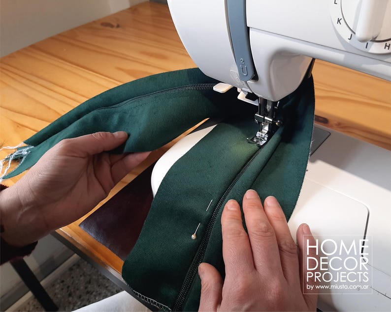 May include: A sewing machine stitching a dark green zipper onto fabric. The fabric is held in place with pins. The machine is white, and the work surface is wood. The image includes the text "HOME DECOR PROJECTS by www.musta.com.ar".