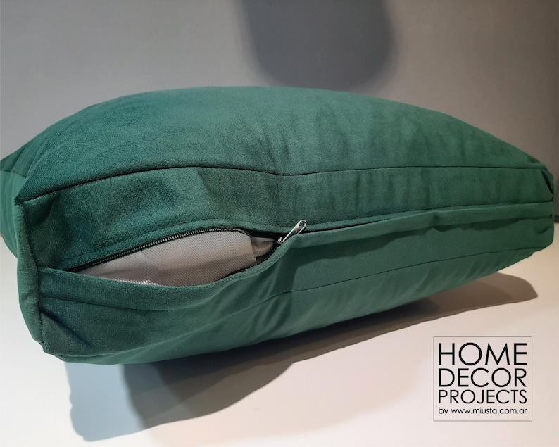 May include: A dark green velvet pillow with a zipper detail. The pillow is rectangular and has a soft, plush appearance. The zipper is partially open, revealing a light gray interior. Home decor project logo in the bottom right corner.