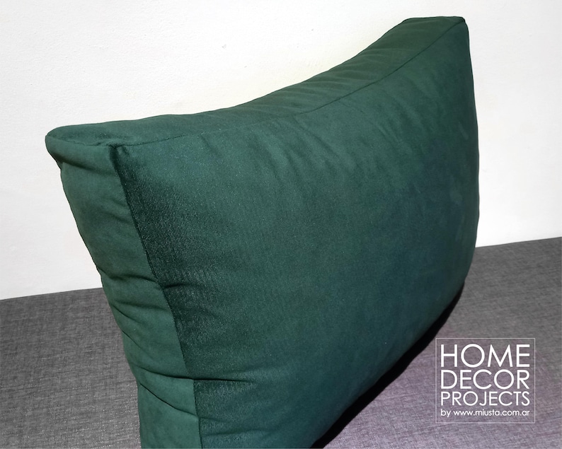 May include: A large, dark green decorative pillow with a triangular shape. The pillow is made of a soft, textured fabric and is designed for home decor. The pillow is placed on a gray surface.