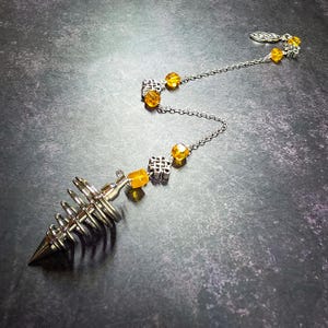May include: A silver-toned metal pendulum with a spiral cone shape attached to a chain. The chain features alternating silver-toned metal knot charms and translucent amber-colored square beads, ending with a small silver-toned leaf-shaped clasp.