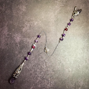 May include: A silver-toned metal chain pendulum featuring a series of purple and deep red beads. The chain includes a central spiral charm and terminates in a pointed, faceted purple crystal encased within an ornate, filigree-style metal cone.