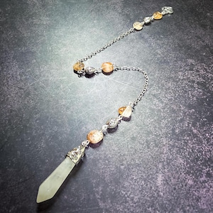 May include: A silver-toned metal chain pendulum featuring a pale green, faceted crystal point at the base. The chain is accented with several round, translucent orange-yellow beads and small, textured silver-toned metal spacer beads throughout.