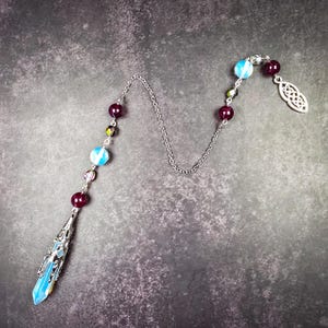 May include: A pendulum featuring a pointed blue crystal encased in a silver-toned metal cap, connected to a silver chain. The chain is adorned with alternating dark red beads, iridescent light blue beads, and a silver-toned Celtic knot charm.