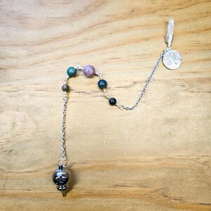 May include: A silver chain pendulum with a metal point and a silver charm with a tree design. The chain has several green, brown, and pink beads.