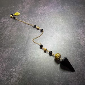 May include: A pendulum featuring a pointed black stone weight attached to a gold-toned chain. The chain is decorated with alternating black beads and ornate gold-toned metal beads, ending with a circular gold-toned charm depicting a tree.