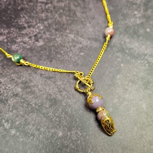 May include: A gold-colored necklace with a pendant featuring purple and tan beads. The pendant has a decorative gold filigree cage at the bottom. The necklace chain has small green and pink beads. The background is a dark gray.