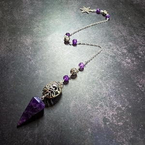 May include: A pendulum featuring a pointed purple amethyst crystal attached to a silver-toned metal chain. The chain is decorated with round purple amethyst beads, ornate silver-toned filigree beads, and a small silver-toned star charm.