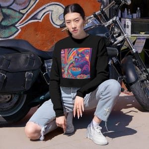 May include: Black cropped sweatshirt featuring a vibrant, colorful graphic of a cat eating from a bowl. The cat is depicted in a psychedelic style with rainbow hues. The sweatshirt is paired with light-wash jeans and white sneakers.