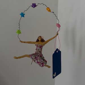 Puede incluir: Una figura caprichosa de una mujer con un vestido floral, a mitad de salto, suspendida de un aro de alambre adornado con estrellas y formas geométricas de colores. Una etiqueta azul cuelga debajo. La figura mide aproximadamente 15 cm de alto.