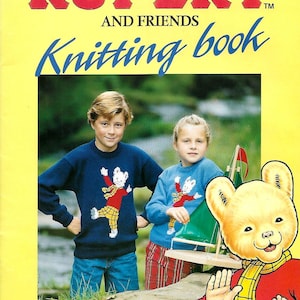 Può includere: La copertina del libro di maglia "The Rupert and Friends Knitting Book" presenta uno sfondo giallo con il titolo in rosso e blu. Due bambini indossano maglioni blu con un disegno di orso. Illustrazione di Rupert l'Orso in basso a destra.
