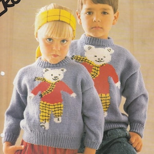 Può includere: Due bambini con maglioni di maglia blu chiaro abbinati con un disegno di orsacchiotto. L'orso ha un corpo rosso, pantaloni a quadri gialli e una sciarpa gialla. Il testo "DOUBLE KNITTING 56-76 cm 22-30 in Five sizes" è visibile.