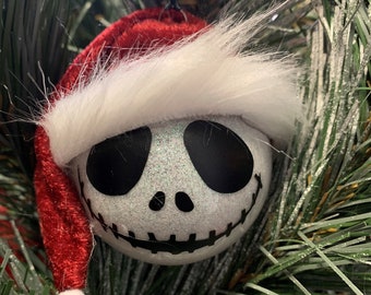 May include: A white Christmas ornament with a black and white Jack Skellington face. The ornament is wearing a red Santa hat with white fur trim.