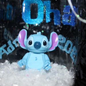Pu&ograve; includere: Un ornamento di vetro trasparente con una figura di Stitch blu chiaro all'interno, circondata da neve finta bianca. L'ornamento presenta un testo glitterato blu che recita "Oh" e "Adrian 2023". La figura di Stitch ha grandi orecchie e un naso blu.