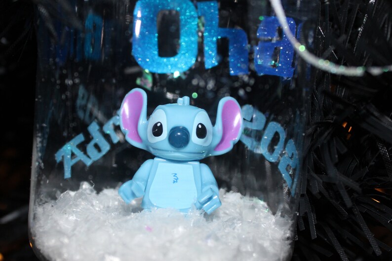 Pu&ograve; includere: Un ornamento di vetro trasparente con una figura di Stitch blu chiaro all'interno, circondata da neve finta bianca. L'ornamento presenta un testo glitterato blu che recita "Oh" e "Adrian 2023". La figura di Stitch ha grandi orecchie e un naso blu.