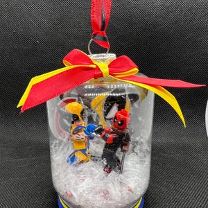 May include: A clear glass snow globe ornament with a red and yellow ribbon bow. Inside the globe are two Lego minifigures, one in a yellow and blue costume and the other in a red and black costume. The globe is filled with white glitter.