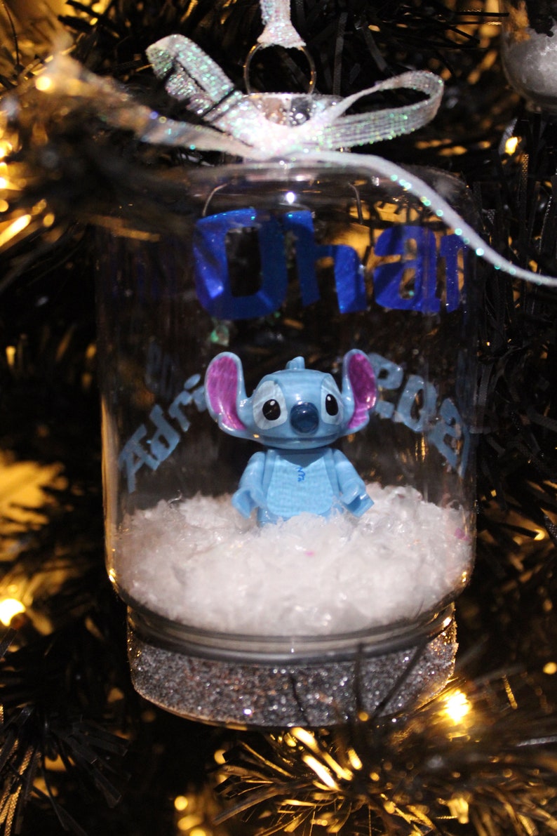 Pu&ograve; includere: Un ornamento di vetro trasparente con una statuina blu di Stitch all'interno, appoggiata su soffice cotone bianco. L'ornamento presenta il testo blu "Ohana" e "Adrian 2023". La base &egrave; glitterata argentata e appesa a un nastro argentato.