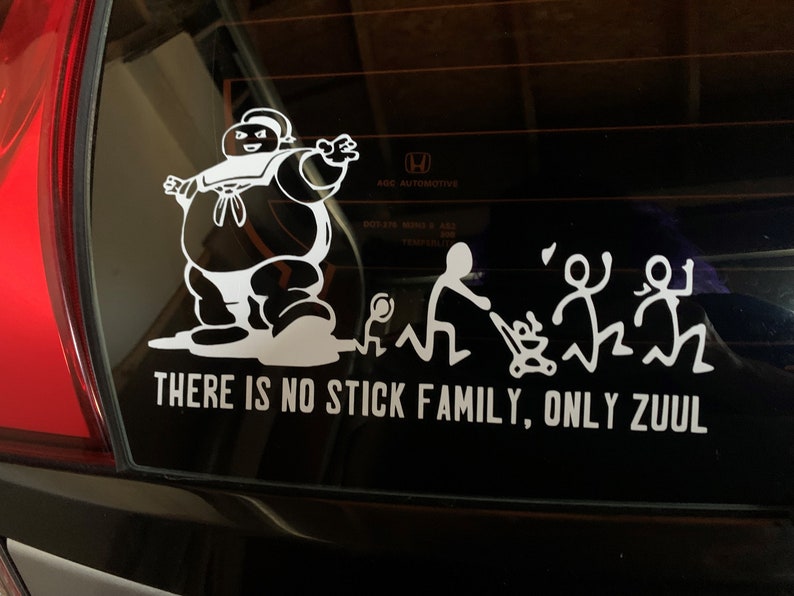 May include: A white vinyl sticker on a black car window. The sticker features a cartoon character resembling the Stay Puft Marshmallow Man from the movie Ghostbusters. The character is walking towards a stick figure family with a baby in a stroller. The text below the image reads "There is no stick family, only Zuul."