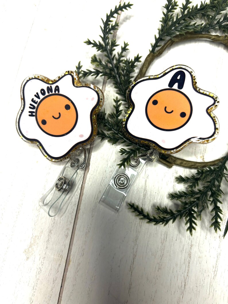 May include: Two gold glitter egg-shaped badge holders with clear plastic straps. One badge holder has the word "HUEVONA" written on it and the other has the letter "A". Both badge holders have a cartoon smiley face egg yolk.