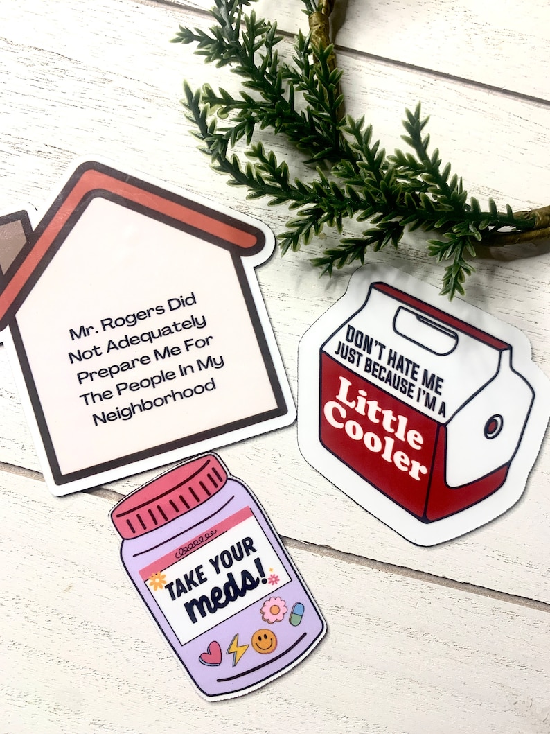 May include: Three stickers with humorous sayings. The first sticker is a house with the text "Mr. Rogers Did Not Adequately Prepare Me For The People In My Neighborhood". The second sticker is a red and white cooler with the text "Don't Hate Me Just Because I'm A Little Cooler". The third sticker is a purple pill bottle with the text "Take Your Meds!" and a heart, lightning bolt, and smiley face.