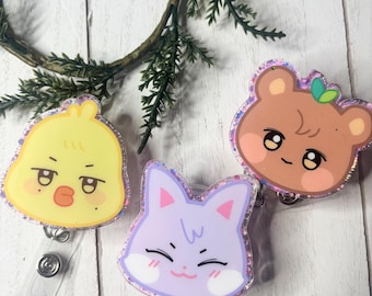 May include: Three cartoon character badge reels. The first is a yellow chick with a grumpy face. The second is a purple cat with a smiling face. The third is a brown bear with a happy face. Each has a glittery border.