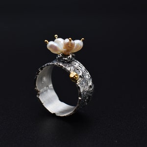 May include: A silver ring with a textured band and a cluster of white pearls on top. The ring has a gold accent on the side.