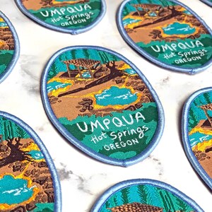 Puede incluir: Una colección de parches bordados con una ilustración escénica de una fuente termal natural con un refugio de madera. Los parches son de forma ovalada con un borde azul y el texto "UMPQUA Hot Springs OREGON" está impreso en cada parche.
