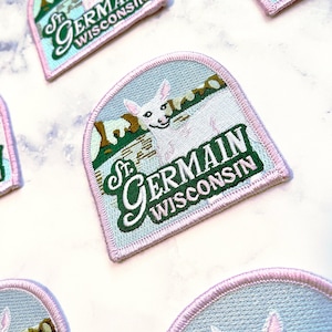 May include: Embroidered patch featuring a white rabbit and the text "St. Germain Wisconsin" in a stylized font. The patch has a light blue background with green and brown accents, and a pink border. Patches are arranged on a white surface.