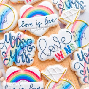 Puede incluir: Surtido de galletas de azúcar decoradas sobre una tabla de madera. Las galletas incluyen diseños de arcoíris, el texto "love is love", formas de anillos de diamantes y las frases "Mrs & Mrs" y "Love Wins".