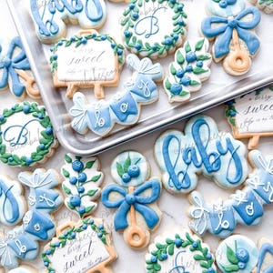 Puede incluir: Una variedad de galletas con temática de bebé, glaseadas en tonos azules y blancos. Las galletas incluyen la palabra "baby", lazos, sonajeros y letreros con texto. Algunas galletas tienen un diseño de arándanos y hojas. Las galletas están dispuestas en una bandeja de metal.