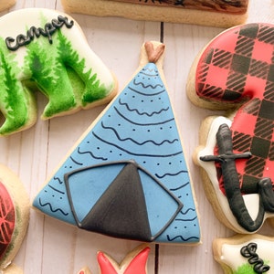 Puede incluir: Una galleta de az&uacute;car decorada en azul y negro con forma de tienda de campa&ntilde;a. La tienda de campa&ntilde;a tiene una solapa negra y puntos blancos y l&iacute;neas onduladas en la tela azul.