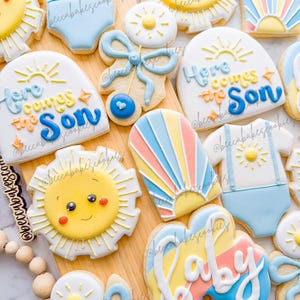 Puede incluir: Surtido de galletas de azúcar decoradas sobre una tabla de madera. Las galletas presentan diseños como un sol sonriente, un body de bebé, un lazo y la frase "Here Comes the Son". Los colores incluyen amarillo, azul, naranja y blanco. El tema es baby shower o bebé recién nacido.