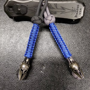 Peut inclure: Deux porte-clés en paracorde bleu et noir avec un charme de casque spartiate argenté. Les porte-clés sont attachés à un étui à couteau noir avec le texte "PIRANHA USA".