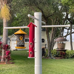 May include: A white PVC bird feeder pole with four bird feeders. The feeders are a yellow hopper feeder, a red tube feeder, a clear tube feeder, and a clear dome feeder. The feeders are filled with birdseed.