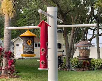 May include: A white PVC pole bird feeder station featuring four hanging feeders. The feeders include a yellow metal mesh feeder, a tall red tube feeder, a clear plastic tube feeder, and a decorative brown dome-topped feeder filled with birdseed.