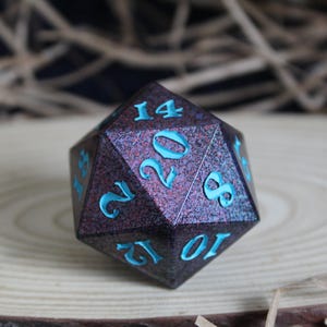 May include: A large, multi-sided die with a dark, textured surface and teal-colored numbers. The die is a deep purple with hints of blue and sits on a wooden surface. The numbers are clearly visible, making it suitable for tabletop games.