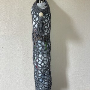 May include: A gray crocheted bag with a wooden bead and tassel hanging from a hook. The bag is filled with plastic grocery bags.
