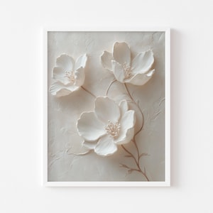 May include: A framed artwork featuring three white, textured flowers with detailed petals and delicate stamens. The flowers are set against a textured, off-white background, with subtle brown stems. The artwork is encased in a white frame.