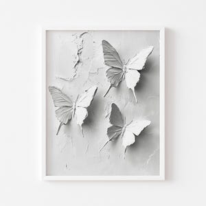 May include: A framed artwork featuring three white butterfly sculptures against a textured, light grey background. The butterflies are arranged in a visually appealing composition, adding a touch of nature-inspired art to any space.