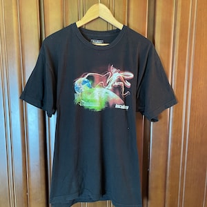 Puede incluir: Camiseta negra con un estampado gráfico de una criatura con alas brillantes y un globo terráqueo. El texto "Incubus" está impreso en la parte inferior del gráfico.