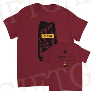May include: A maroon t-shirt featuring a black graphic and the text "R.E.M." in a yellow rectangle. The back of the shirt displays handwritten text and a question mark, with the band's name again. A casual, music-themed apparel item.