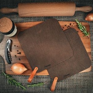 Könnte beinhalten: Draufsicht auf ein Holzschneidebrett mit zwei braunen Lederuntersetzern, einem Nudelholz, einem Glas, einem Korkenzieher, einem Messer und Rosmarinzweigen. Die Untersetzer haben Ledergriffe.