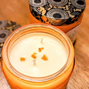 May include: A jar candle with a white wax interior and three wicks. The candle is in an orange glass jar with a decorative pattern. The lid is black with a floral design. The text "Pumpkin Spice" is at the bottom.