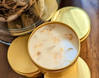 May include: A set of four gold candle tins with a white candle inside each tin. The candles have small pieces of dried flowers embedded in the wax.