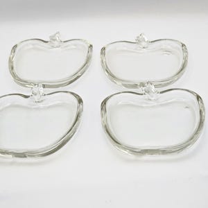 May include: Four clear glass apple-shaped dishes. Each dish has a stem detail at the top. The dishes are arranged in a square formation against a white background. The glass is transparent, allowing light to pass through.