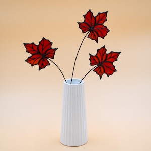 May include: Three red stained glass maple leaves in a white vase with vertical ridges. The leaves are on black wire stems.