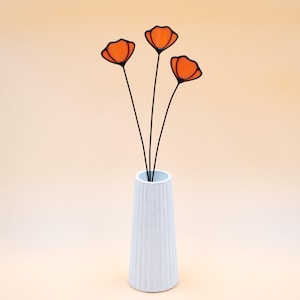 May include: A white, ribbed vase holds three orange and black floral stems. The flowers have a stained glass appearance, with black outlines and orange petals. The stems are thin and black, creating a minimalist, modern aesthetic.