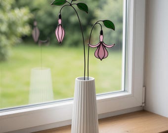 May include: Two stained glass fuchsia flowers with pink petals and green leaves on thin black metal stems, displayed in a white ribbed ceramic vase. The flowers feature delicate, teardrop-shaped pink blossoms and dark green foliage.