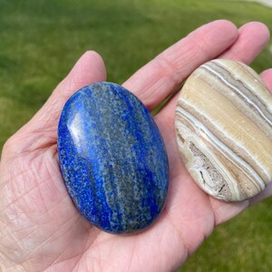 May include: Two polished oval-shaped stones. One is a blue lapis lazuli stone with white and gold veins. The other is a light brown and white banded stone.
