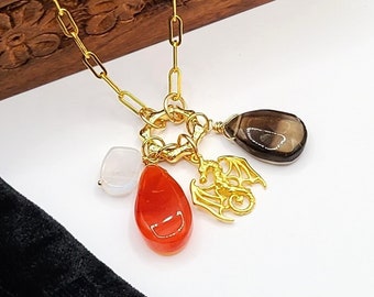Pode incluir: Colar com pingentes dourado com corrente de clip e vários pingentes. Os pingentes incluem uma pedra laranja em forma de gota, um dragão, um quartzo fumê e uma pedra branca. O colar é exibido em uma superfície branca.